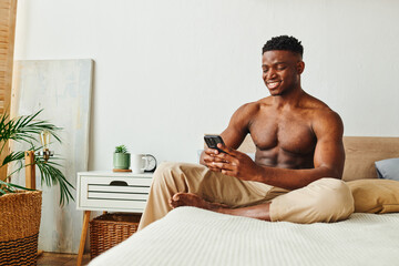 joyous and muscular african american in pajama pants browsing social media om smartphone in bedroom