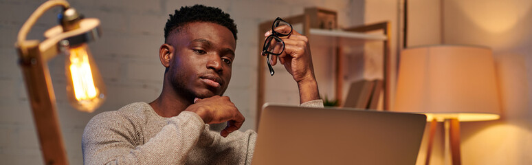 pensive african american freelancer holding eyeglasses looking at laptop in home office, banner