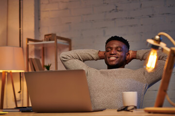 pleased and relaxed african american man looking at laptop in home office at night, freelancer