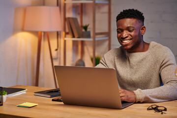 cheerful african american man working on laptop in home office at night, independent freelancer