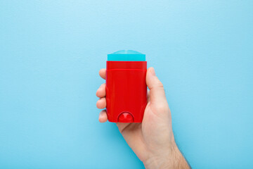 Young adult man hand holding and showing red plastic stick of armpit deodorant on light blue table background. Pastel color. Male daily beauty product. Closeup. Top down view. - obrazy, fototapety, plakaty