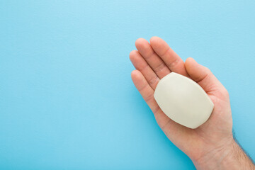 Young adult man opened palm holding gray natural soap on blue table background. Pastel color. Care about clean and soft body skin. Daily beauty product. Closeup. Empty place for text. Top down view. - obrazy, fototapety, plakaty