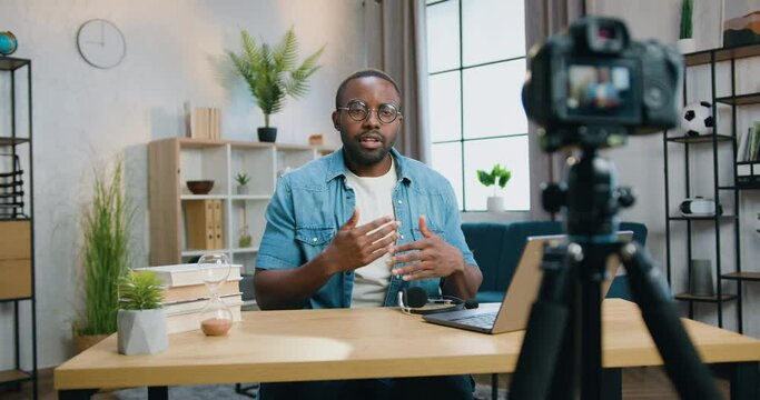 Attractive african man blogger in glasses filming new video for his audience with professional videocamera on home interior background