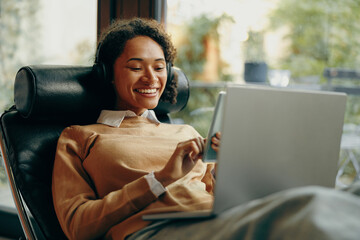 Smiling woman in headphones is use smartphone while sitting home on chair with laptop  - Powered by Adobe