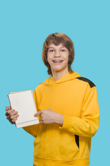 Young smiling teen boy holding notebook isolated on blue background, Teen boy's bright smile radiates as he holds a notebook with confidence.