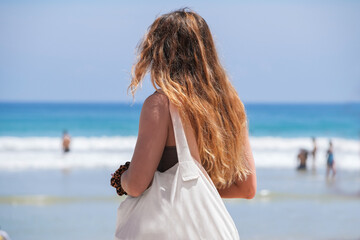 Fototapeta na wymiar A girl with long hair stands with her back against the background of the ocean on the beach on a sunny day