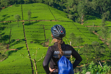 Blonde hair female cyclist in helmet looking at tea plantations and forest, rear view