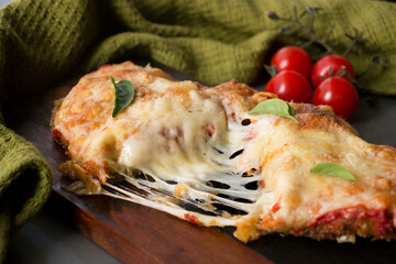 Breaded meat fillet with tomato sauce, melted cheese and oregano, on a wooden board, in the background tomatoes and a green cloth for production