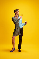 Young male model half-wearing in woman's and man's clothes posing holding glasses of champagne and...