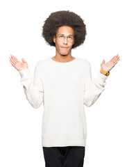 Young african american man with afro hair wearing glasses clueless and confused expression with arms and hands raised. Doubt concept.