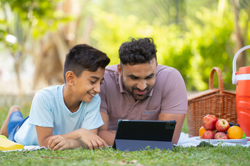 Happy smiling indian father with son using digital tablet during weekend picnic at park - concept of cyberspace, family fun and Joyful interaction - obrazy, fototapety, plakaty