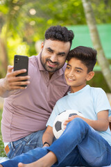Vertical shot of cheerful indian father with son taking selfie on mobile phone at park - concept of social media, family holidays and relationship