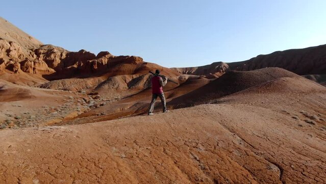 A guy with a camera on a tripod walks along a hill against the backdrop of unusual red rocky mountains. A man with a camera in a red down jacket. Azhirzhar tract, Kazakhstan