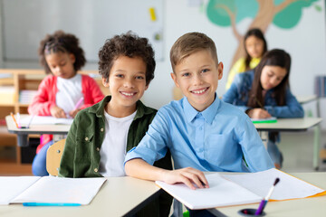 Intercultural schoolkids boys sitting at desk during lesson, looking at camera and smiling