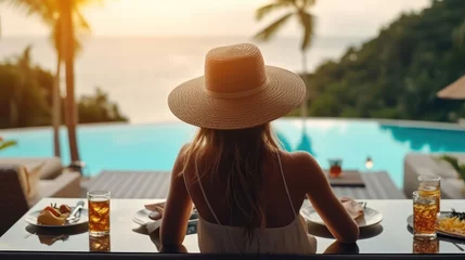 Deurstickers Dinner with sea view in luxury hotel. Woman in straw hat near swimming pool, eating food and enjoy ocean view. Dinner table on tropical vacation. Back view. Concept of travel, holidays, weekend. © Santy Hong