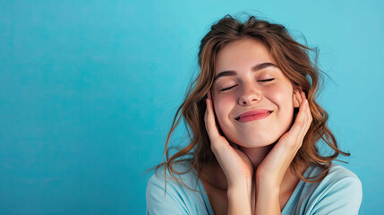 Cheerful female smiling for camera and clasping hands under cheek while pretending to sleep against blue background - obrazy, fototapety, plakaty