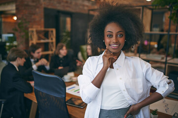 African-American woman smiling at camera while classmates studying with laptop on background. Public library. Concept of business, study, co-working space, teamwork, collaboration, classmates.