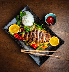 Asian food. Peking duck, rice and vegetables on a black ceramic plate, on the background of a wooden table