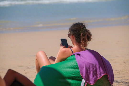 Woman sits on pouf resting and scrolls the feed on her smartphone on the beach 