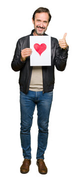 Handsome middle age man holding paper with red heart happy with big smile doing ok sign, thumb up with fingers, excellent sign