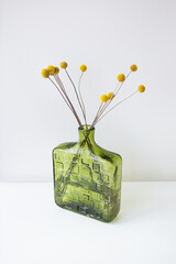 A green textured glass vase with yellow Craspedia flowers against a white background. Simple and...