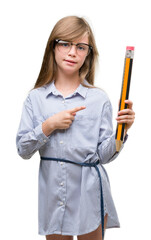 Young blonde child holding big pencil very happy pointing with hand and finger