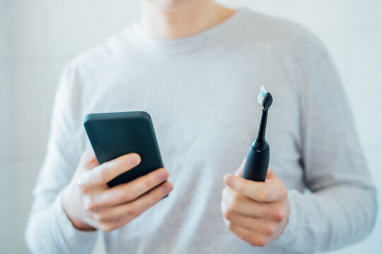 Close up man puts settings of his electronic toothbrush with mobile phone app. Wireless connecting sonic toothbrush with smart phone app. Modern home health care technology concept. Selective focus