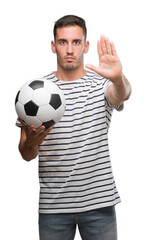 Handsome young man holding soccer football with open hand doing stop sign with serious and confident expression, defense gesture
