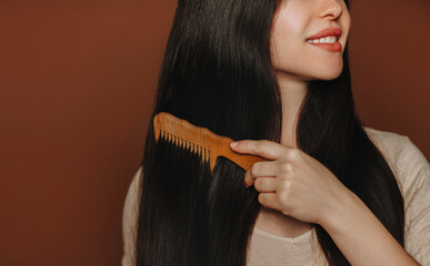 Young happy brunette woman combing her long healthy brown hair and smiling at camera while posing isolated on dark background