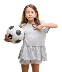 Brunette hispanic girl holding soccer football ball with surprise face pointing finger to himself