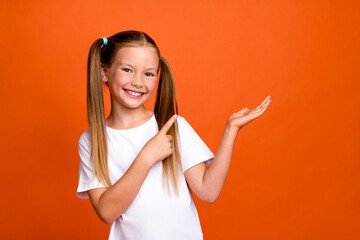 Photo portrait of pretty small girl palm hold point empty space wear trendy white outfit isolated on orange color background