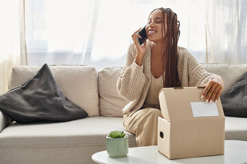 joyful attractive african american woman in comfy homewear talking by phone with hand on parcel