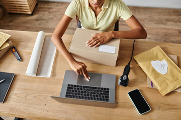 cropped view of young african american female retailer packing box and working on laptop, delivery