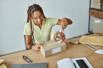 jolly young african american female retailer using tape on cardboard box, delivery concept