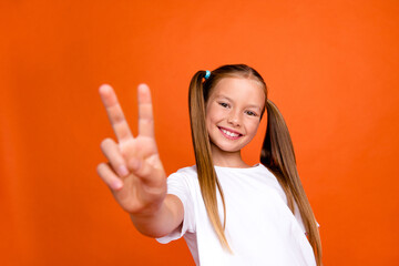 Photo portrait of pretty small girl show v-sign hello gesture wear trendy white outfit isolated on orange color background