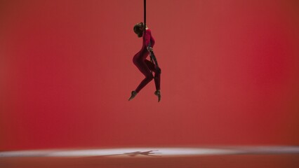 Female gymnast isolated on red studio background. Girl aerial dancer in bodysuit on acrobatic trapeze with straps perform element.