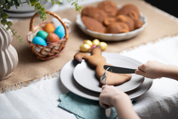 Unrecognizable child cutting gingerbread cookie shaped as Easter bunny with fork and table knife at festive decorated table. Holiday traditions, learning table manners and dining etiquette