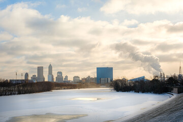 A view of the Indianapolis skyline from White River State Park. The White River is frozen in the foreground with the city in the background. The sky is colorful from the mix of sun, clouds, and sky.
