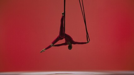Female gymnast isolated on red studio background. Girl aerial dancer in bodysuit on acrobatic trapeze with straps perform element.