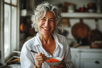 Smiling beautiful elderly woman eating red caviar by the spoon in the kitchen. Concepts: healthy lifestyle, active longevity, proper nutrition, healthy fats, healthy skin - obrazy, fototapety, plakaty