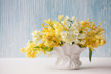 Spring bouquet of yellow hyacinths, daffodils, mimosa in a vase on a table, blue background wall. Closeup, blur, soft focus.