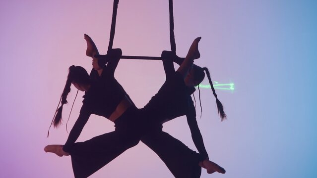 Female gymnastic duet isolated on laser blue neon studio background. Girls aerial dancers flying on gymnastic trapeze with straps.