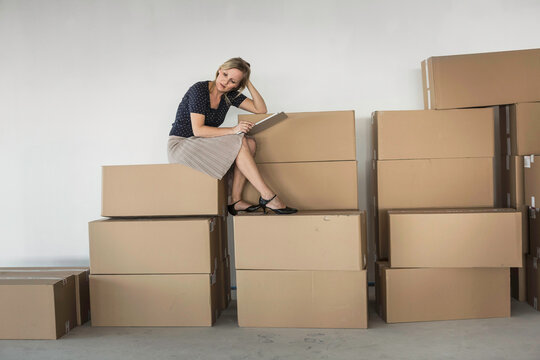 Female owner of a startup sits on top of moving boxes in the office with business plan in hand.