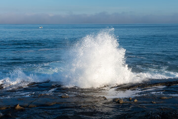 2020-01-03 WATER CRASHING ON THE ROCKY SHORELINE CAUSING SPRAY WITH A CALM PACIFIC OCEAN IN THE BACKGROUND AND A BOAT IN THE BACKGROUND IN LA JOLLA CALIFORNIA