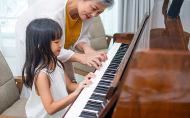 Asian elderly grandmother teaching little sweet girl playing piano, smiling with happiness, staying at cozy home, spending time together. Family, Love, Education Concept.