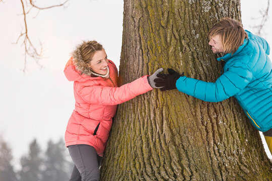 Young beautiful woman and her boyfriend chasing each other around a tree in winter. Wolfratshausen, Bavaria, Germany 