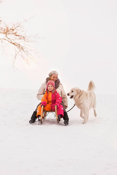 Older woman sledding down a hill with granddaughter and dog in winter. Wolfratshausen, Bavaria, Germany