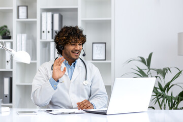 Engaging male Hindu doctor in white coat with headset waving at the camera, providing a warm...