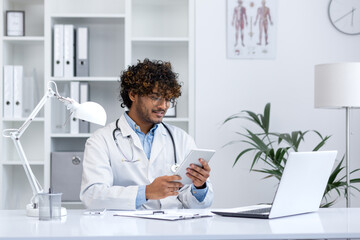 A professional young doctor in a white coat attentively uses a digital tablet in a bright medical...