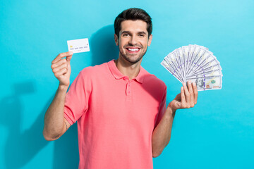 Portrait of young man wearing pink t shirt holding banknotes dollars and cashback credit card isolated on aquamarine color background